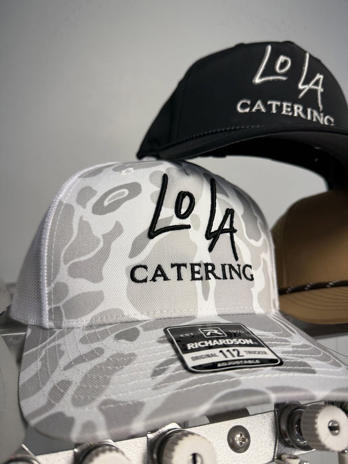 Camo and black trucker hats embroidered with the LoLa Catering logo